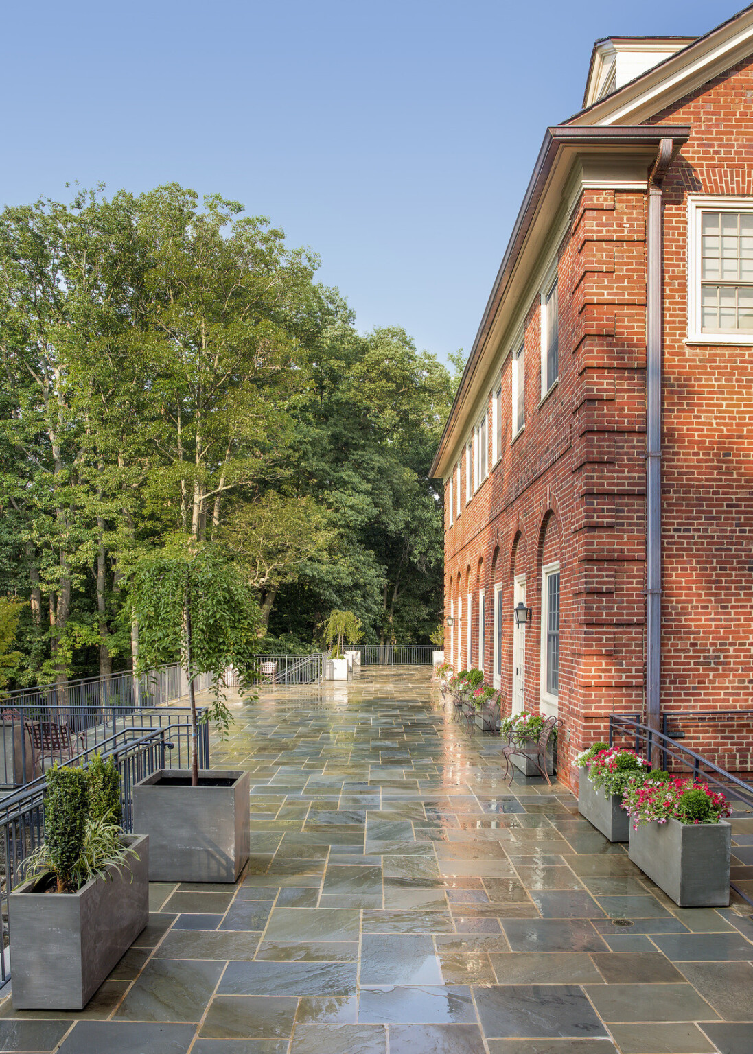 Outdoor view of wet stone walkway, concrete planters with bright flowers, small trees, dormitory building and woodlands in background