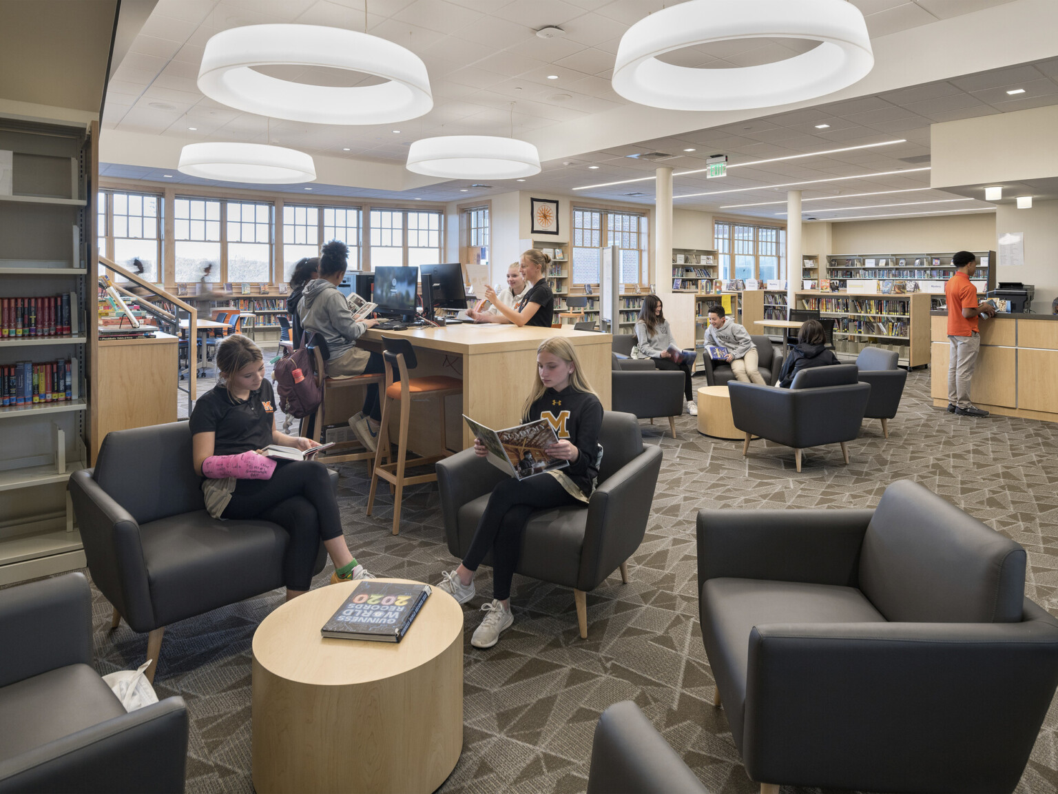 Middle school library with half height bookshelves, flexible tables and seating, arm chairs, light-colored woods, muted grey seating and modern circular lighting suspended from the ceiling