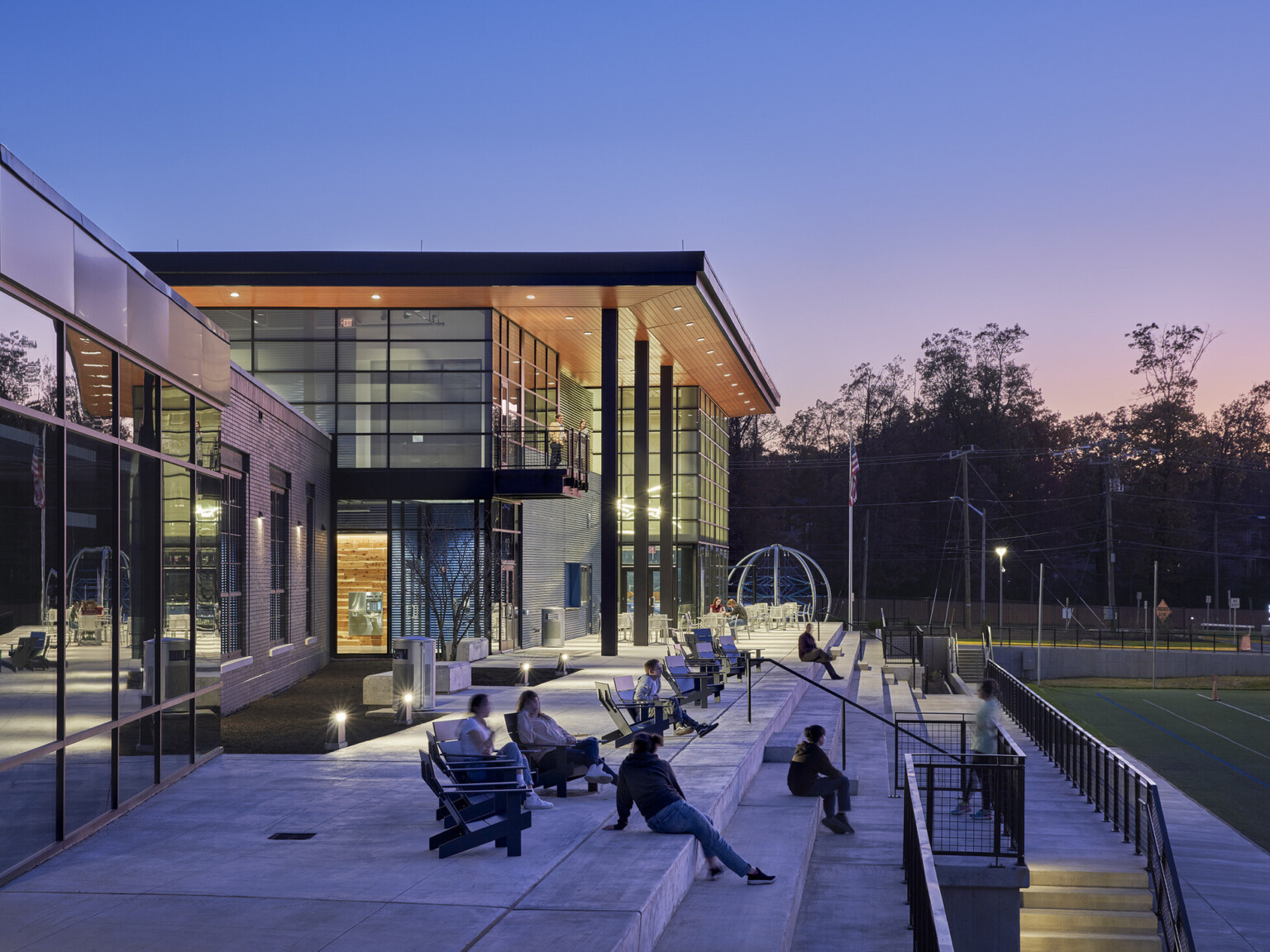 students relaxing on cement patio outside a modern school with glass walls, wood overhang held up with metal columns