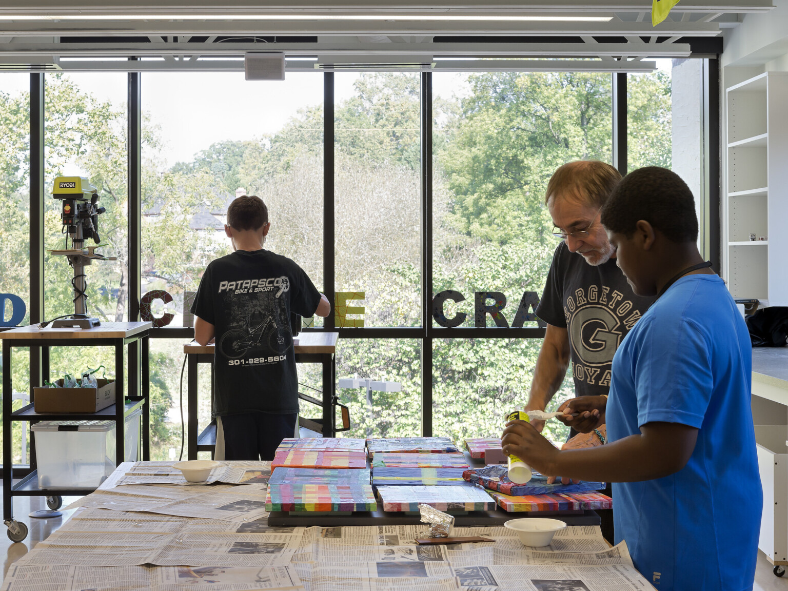 two students with teacher in well-lit open-concept learning environment, floor-ceiling windows, natural lighting and light wood desks and work tables, neon light sign and art supplies