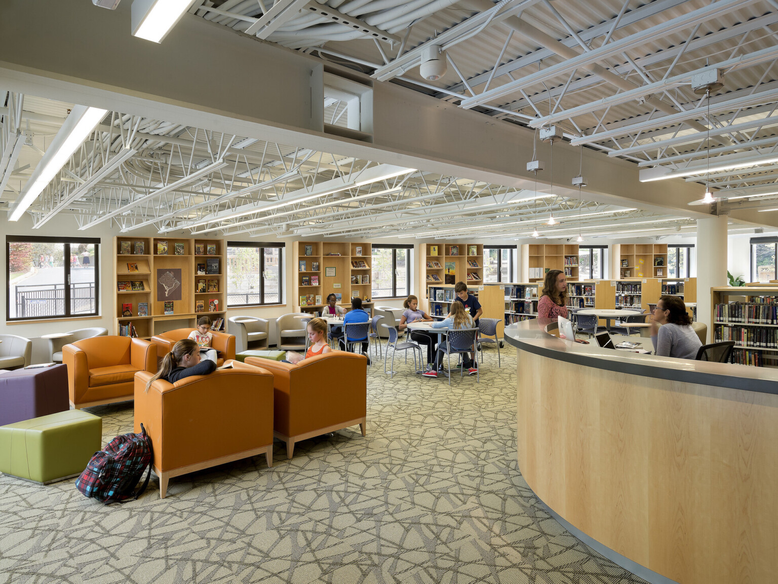 students and librarian in library on laptops among double-height ceilings, a bright, red round rug brings the groups together, natural daylight and skylights, comfy, modern orange leboucle-covered seating