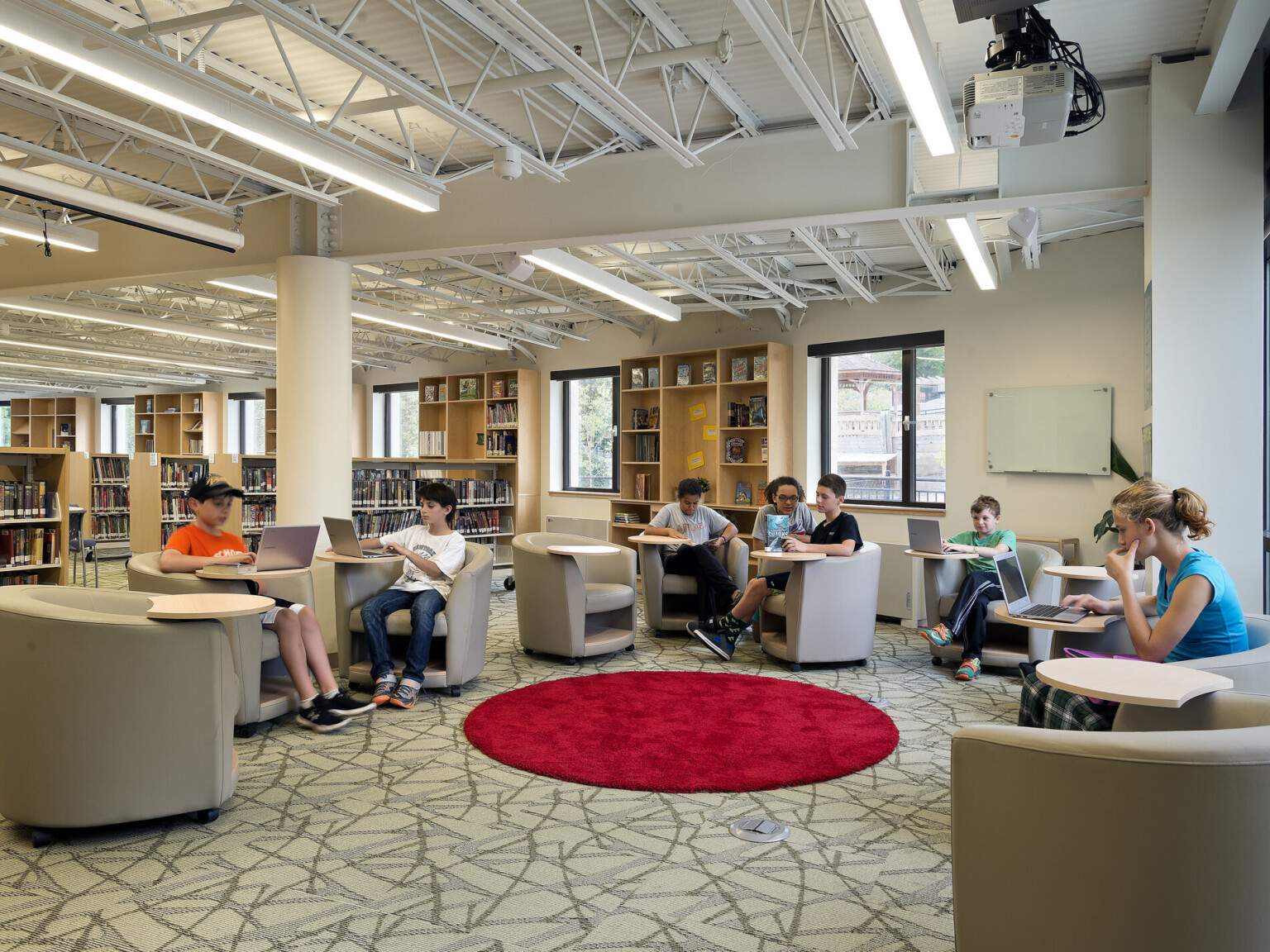 students in library on laptops among double-height ceilings, a bright, red round rug brings the groups together, natural daylight and skylights
