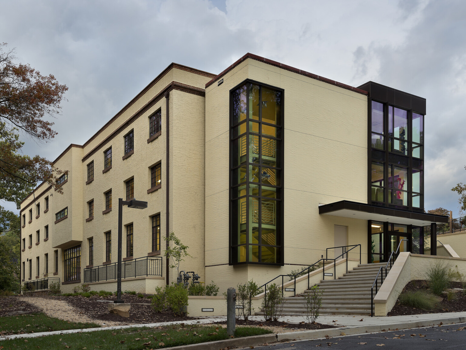 exterior of three-story building, concrete, tile and steel, interior floor-to-ceiling windows, outdoor steel stairway, tall trees and grass below cloudy skies at midday