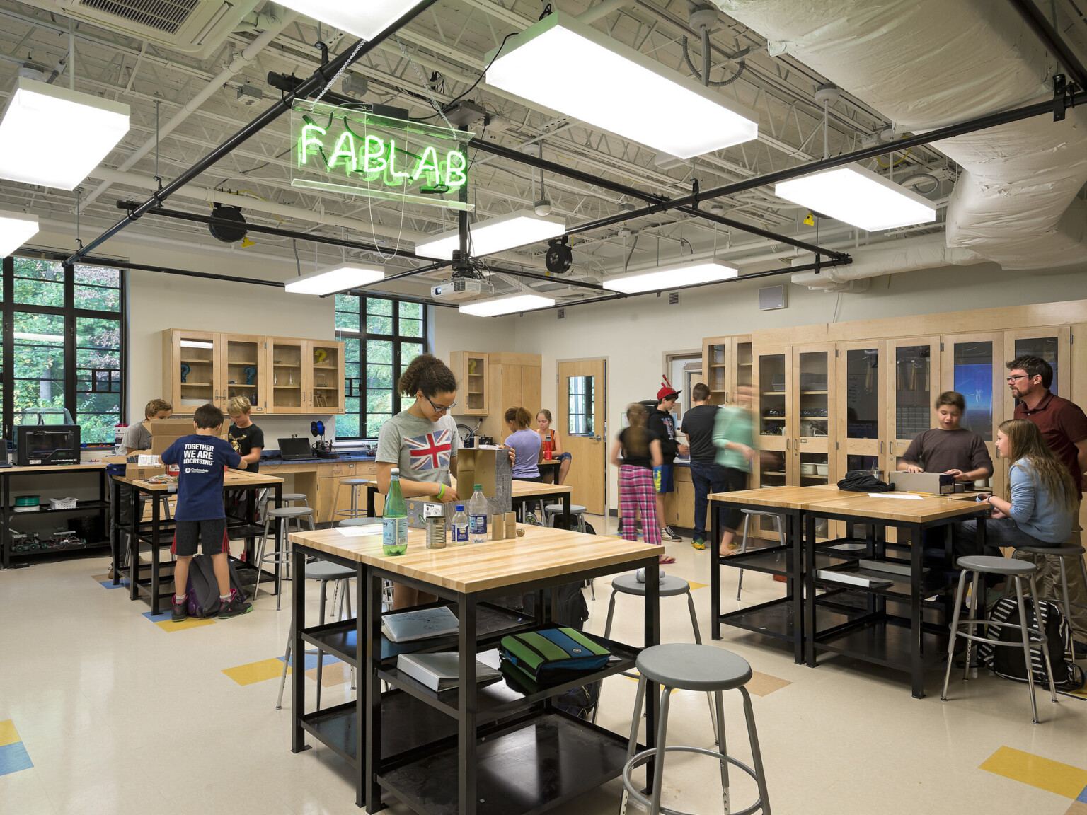 students collaborate in well-lit open-concept learning environment, floor-ceiling windows, natural lighting and natural light wood desks and work tables, neon light sign says, "fablab"