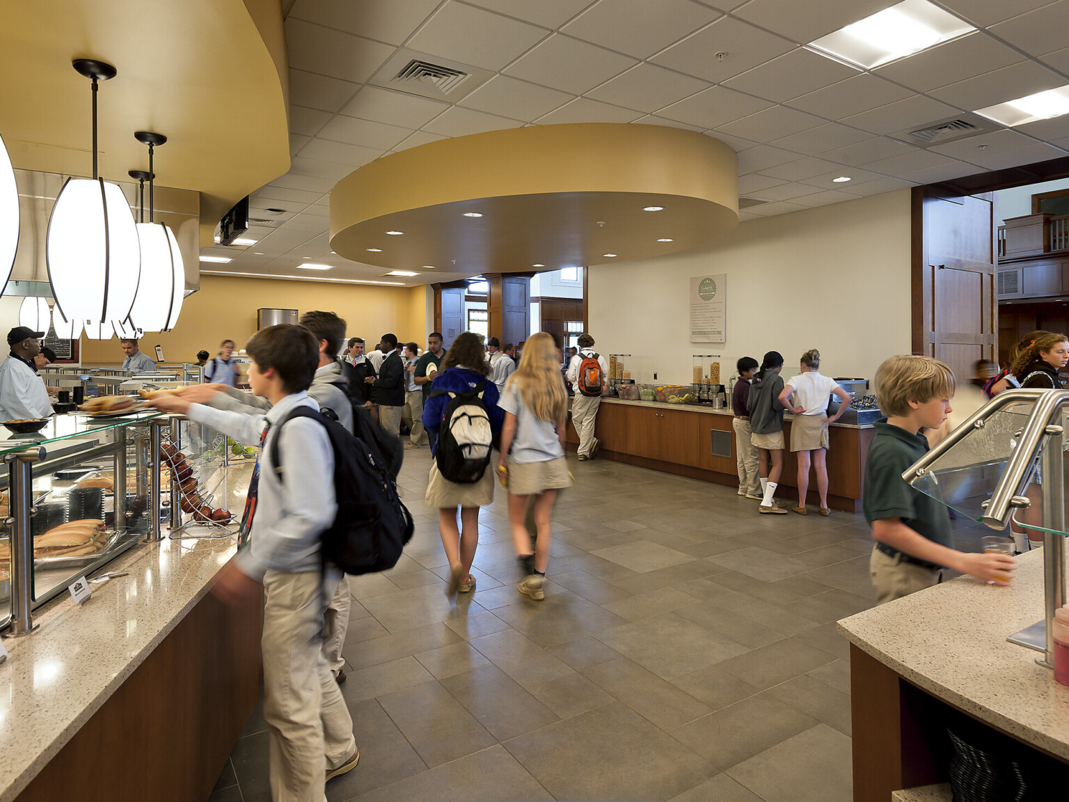 Students enjoy a sunny yellow, modern cafeteria designed for quick, casual dining, custom pendant lights, stone tile flooring