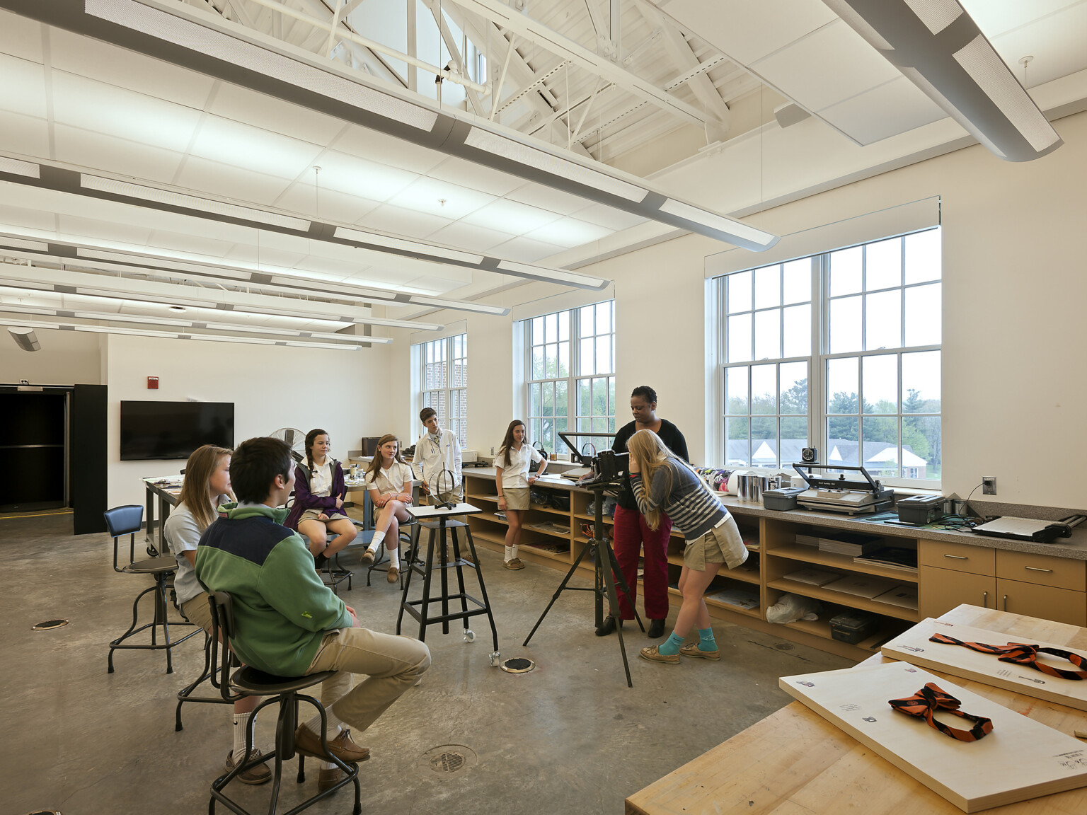 student looks through lens on tripod surrounded by seated classmates and instructor, double-height ceilings, wide floor-to-ceiling windows, concrete polished flooring