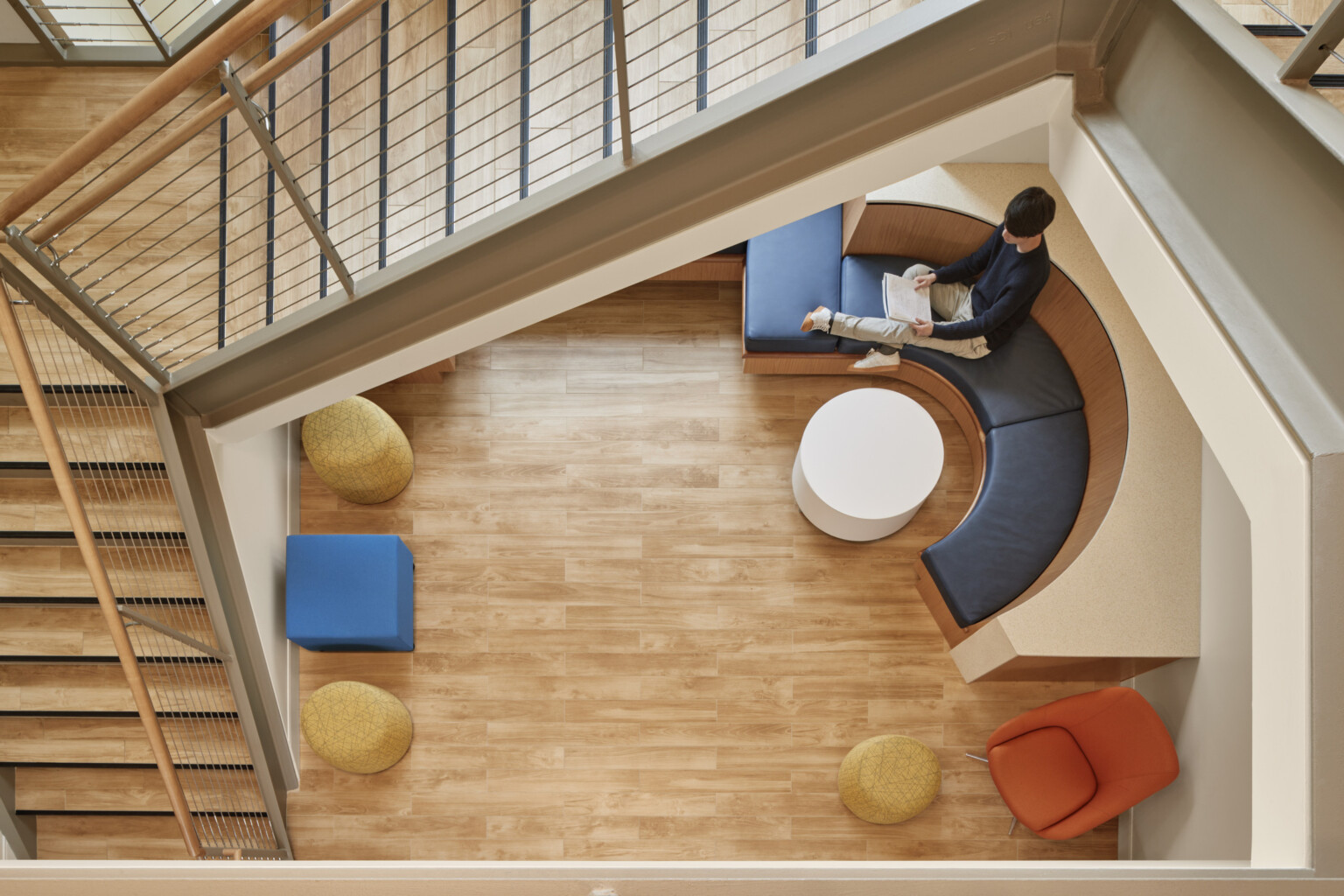 Top of wood stairwell looking to mixed comfortable seating at lower level, rounded bench seat next to window lit with natural light