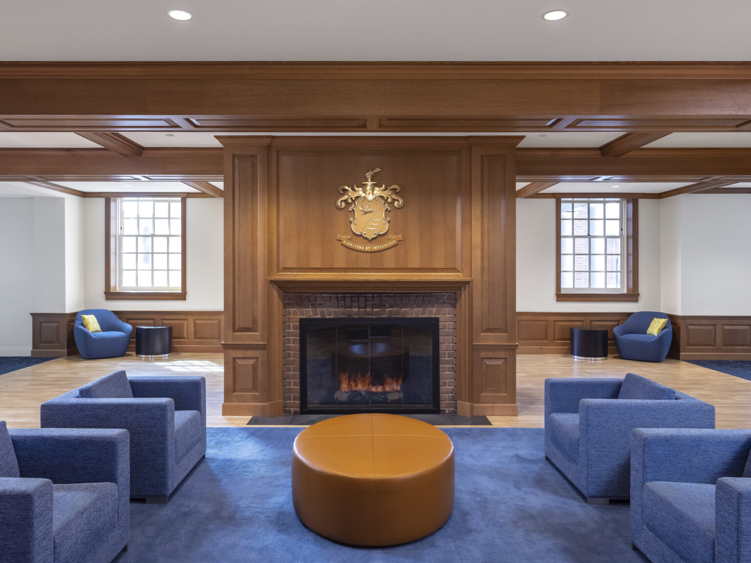 Blue armchair and ottoman in common room seating area in front of fireplace, coat of arms on mantle, large windows, coffered ceiling