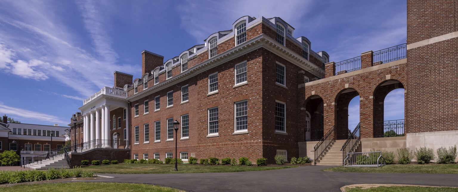 Side view of brick dorm with large windows, connecting to a building by covered outdoor arcade walkway with widows walk above