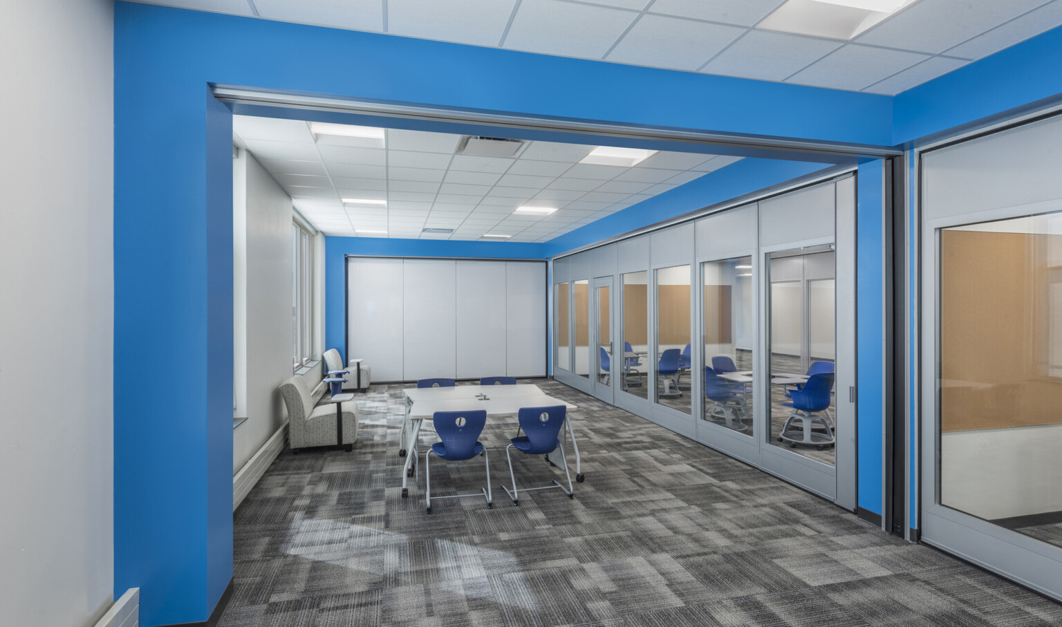 Large whiteboard in blue room with mixed seating. Silver and glass flexible wall, right, with flexible desks in next room