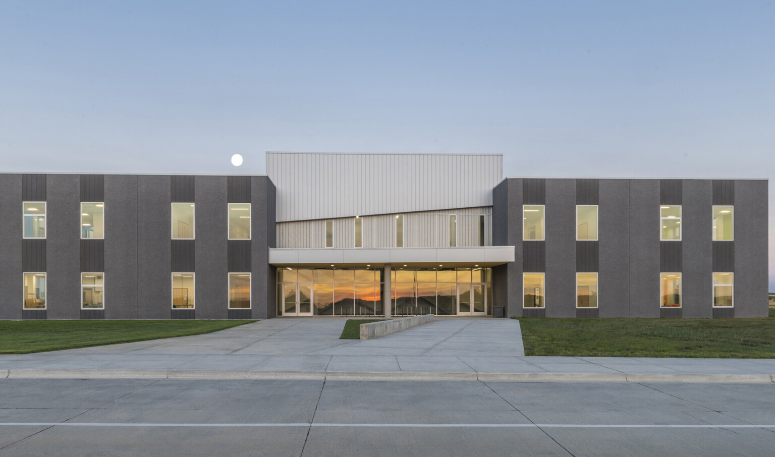 Dark grey building sections frame recessed center with glass base and canopy below silver panel 2nd floor. Driveway to front