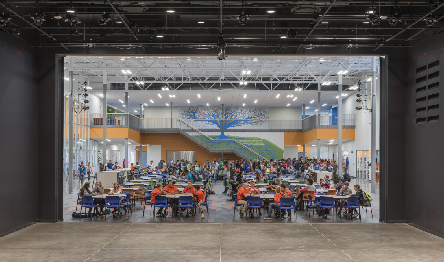 Double height room with tables and chairs on 1st floor. Orange stairs at back wall with abstract blue and orange tree mural
