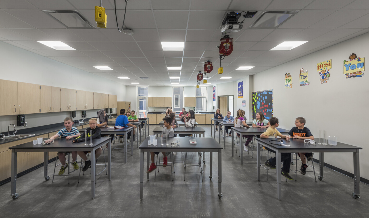 White science lab with light wood cabinets, black counters and rows of lab tables. Extension cords above, windows at back