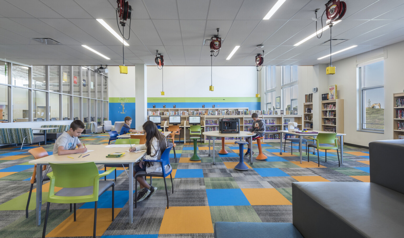 Flexible seating space in carpeted room with floor to ceiling windows to hall, right, and rows of half height book shelves
