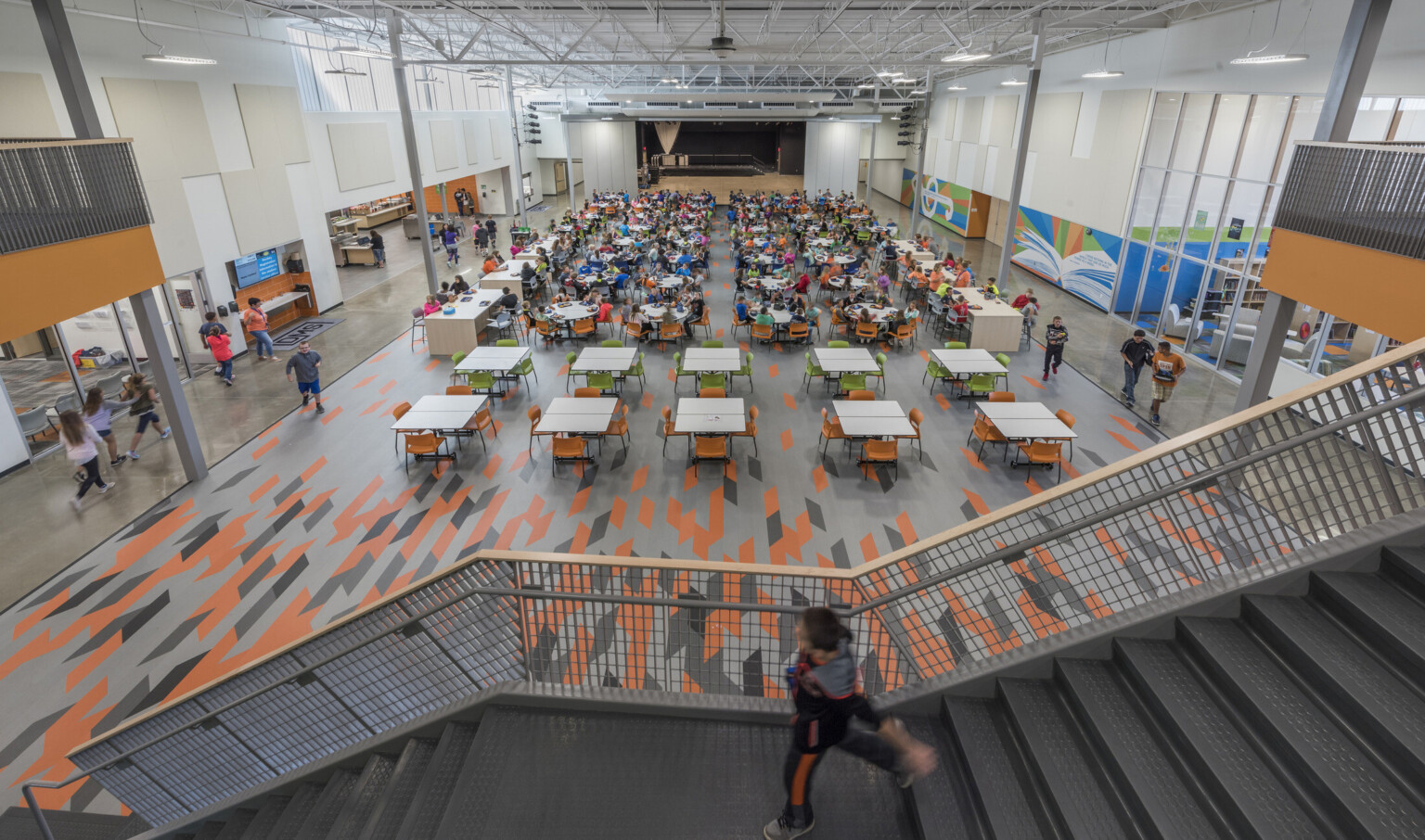 View from stairs down to cafeteria tables. Book and music murals on right by library. Cafeteria counters and sink, left