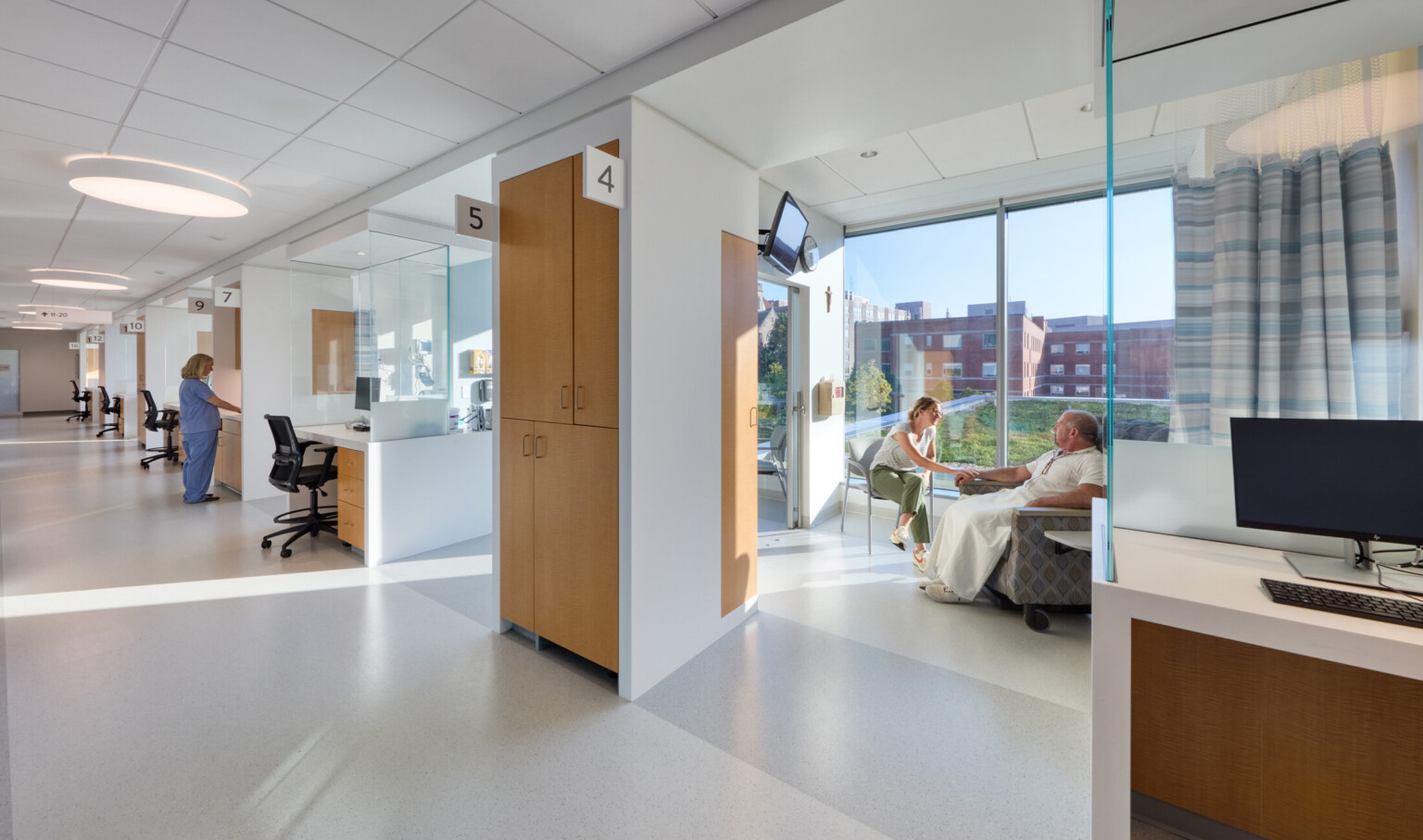 Physician and patient area in the OSF Cancer Institute and Proton Therapy building; bright natural light behind the patients and doctors in the space; modern white with light wood details on the cabinets; computer stations throughout