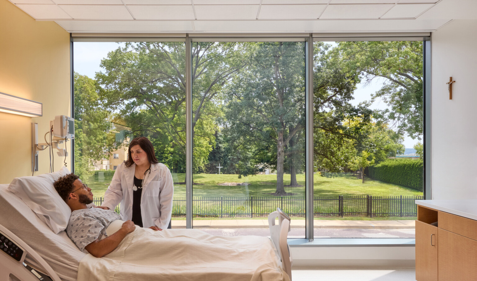 patient room in the OSF Cancer Institute and Proton Therapy building; patient being attended to by a caregiver in a bed; floor to ceiling windows behind offer views of the greenspace and sky outside; machines for measuring vitals on the wall