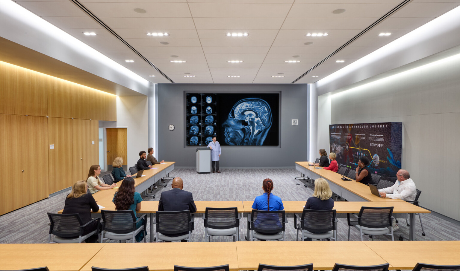 Lecture and gathering space in the OSF Cancer Institute and Proton Therapy building; doctors looking at a large brain scan on the back wall with tables oriented toward a speaker and the scans; LED lights and light wood features make the space bright