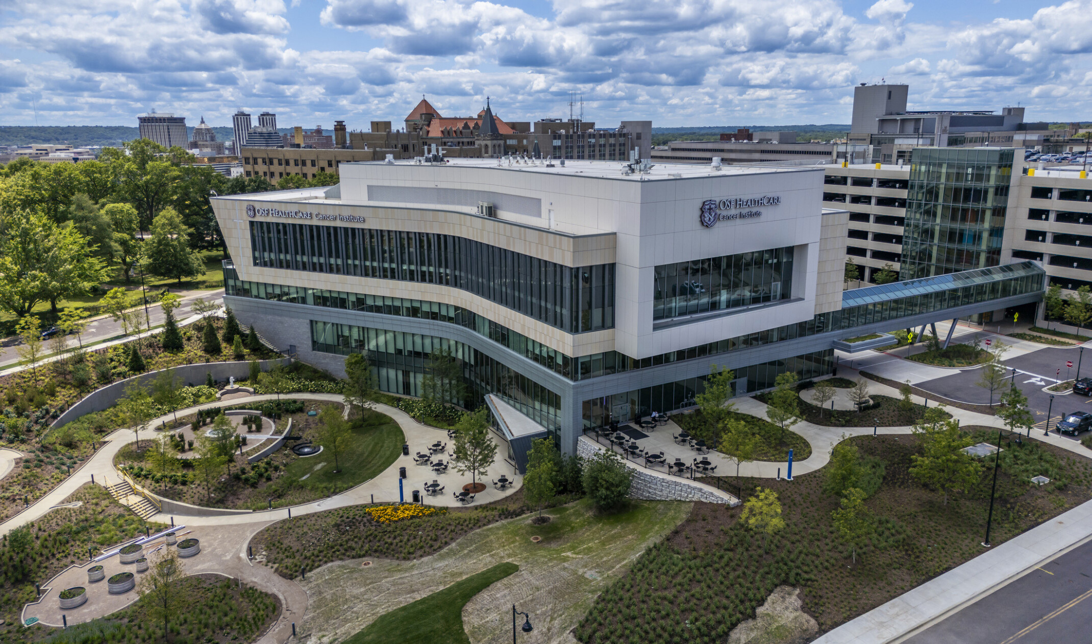 OSF Cancer Institute and Proton Therapy healthcare building; foreground greenery and walking space with the large modern building behind; featuring floor to ceiling windows facing out to the green space