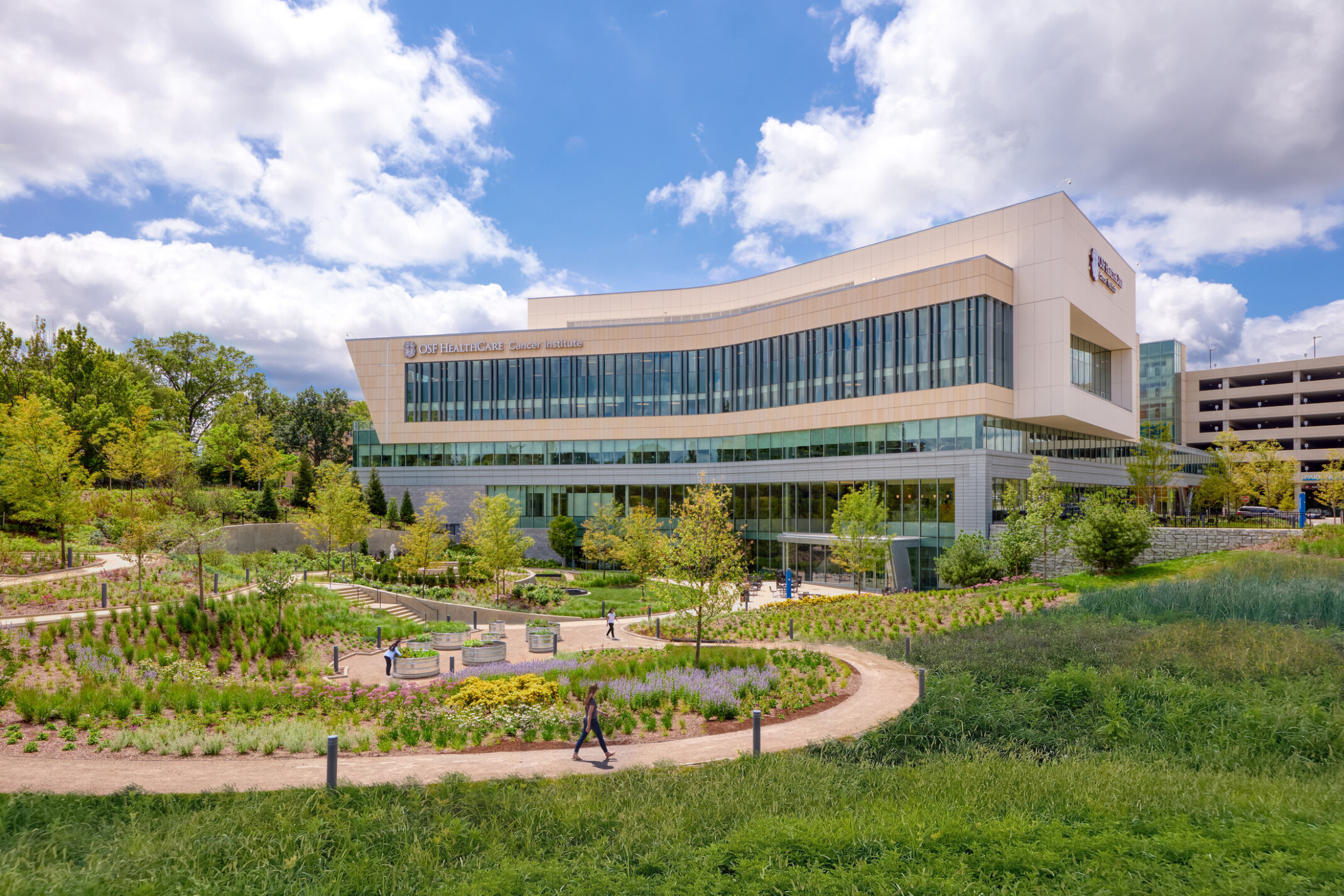 OSF Cancer Institute and Proton Therapy healthcare building; foreground greenery and walking space with the large modern building behind; featuring floor to ceiling windows facing out to the green space OSF Cancer Institute and Proton Therapy healthcare building; foreground greenery and walking space with the large modern building behind; featuring floor to ceiling windows facing out to the green space