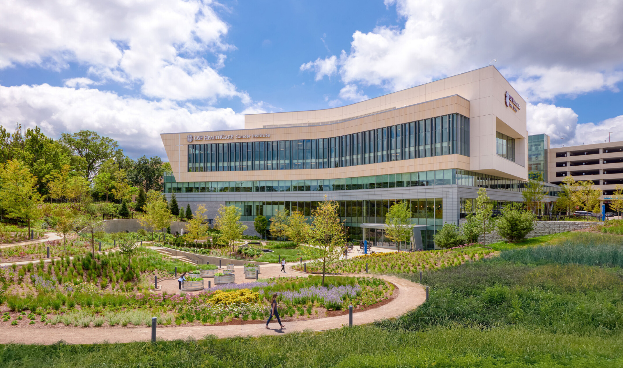 OSF Cancer Institute and Proton Therapy healthcare building; foreground greenery and walking space with the large modern building behind; featuring floor to ceiling windows facing out to the green space