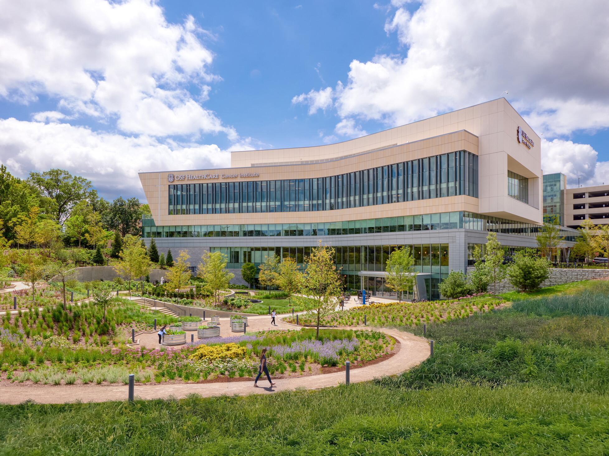 OSF Cancer Institute and Proton Therapy healthcare building; foreground greenery and walking space with the large modern building behind; featuring floor to ceiling windows facing out to the green space