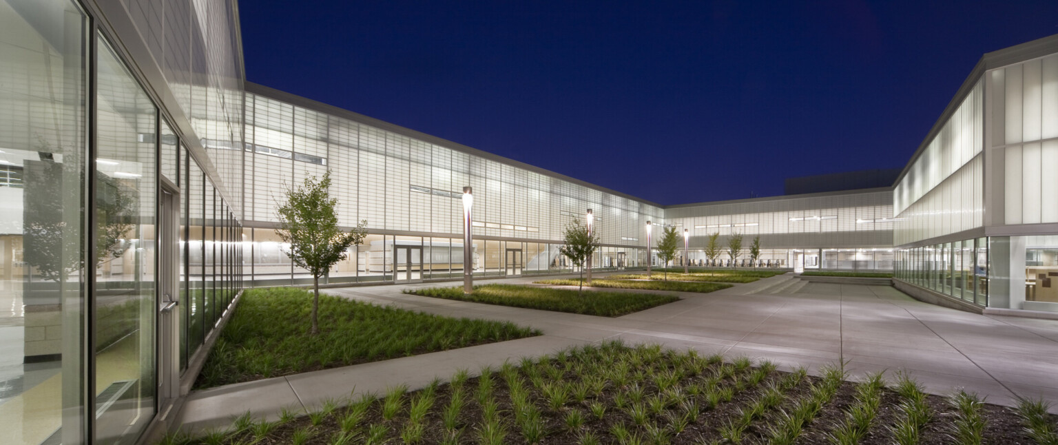 Exterior view of campus, floor-to-ceiling windows, cut out windows, high-ceilings, translucent wall panels filter natural light into the school’s common spaces, these panels shine as a beacon of light on the prairie at night, landscaped courtyards and grounds, trees