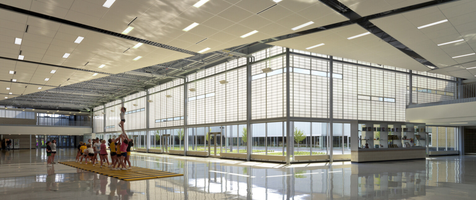 interior view lobby, floor-to-ceiling windows, cut-out windows, high-ceilings, translucent wall panels filter natural light into the school’s common spaces, gleaming, polished concrete floors, cheer team lift cheerleader over their heads on safety mat