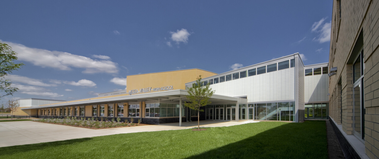 Exterior view of campus main entrance daytime, signage, floor-to-ceiling windows, cut out windows, high-ceilings, translucent wall panels filter natural light into the school’s common spaces, landscaped courtyards and grounds, trees, blue skies