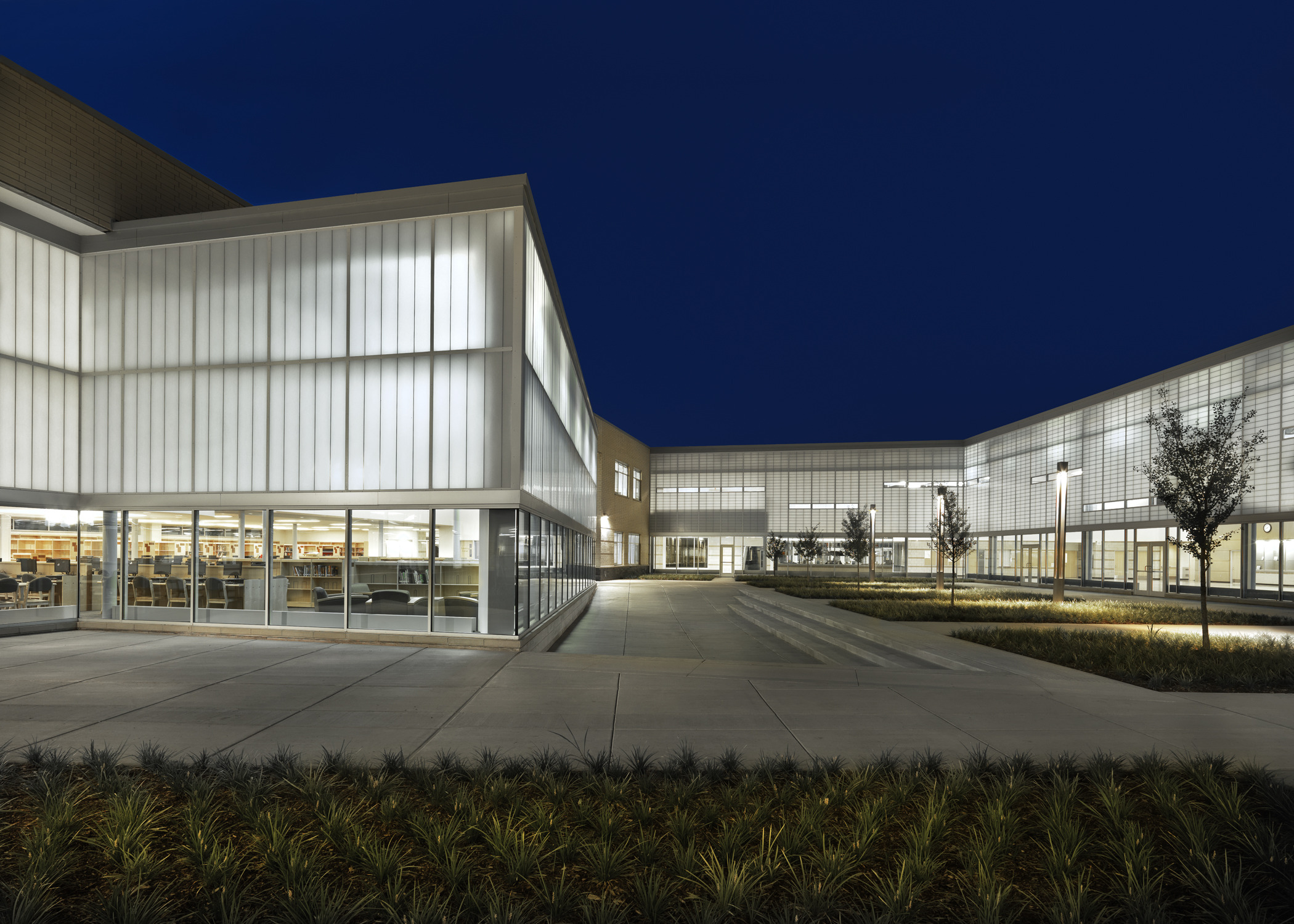 Exterior view of campus, floor-to-ceiling windows, cut out windows, high-ceilings, translucent wall panels filter natural light into the school’s common spaces, these panels shine as a beacon of light on the prairie at night, landscaped courtyards and grounds, trees