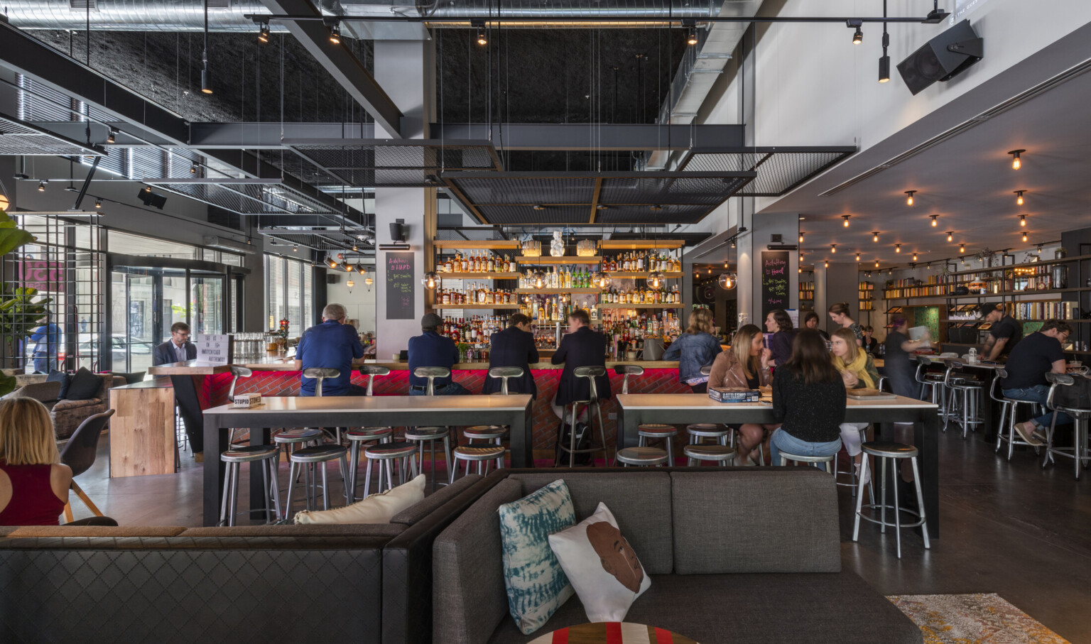 Red brick bar with wood counter and behind the bar shelving. High top tables and stools sit between bar and couches