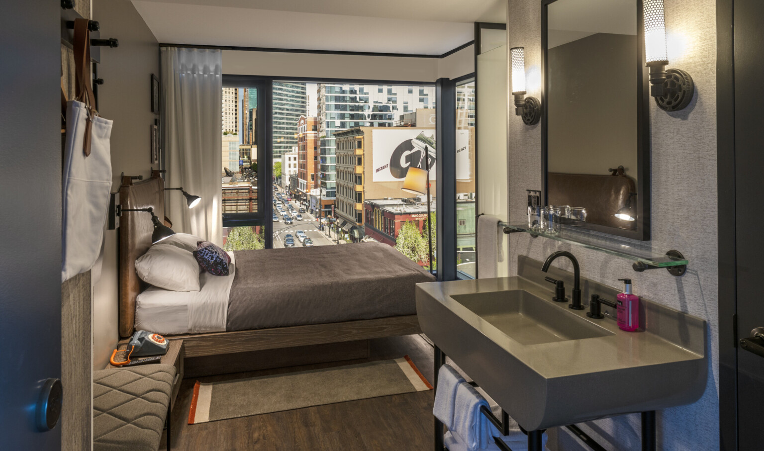 View of guest room from grey sink, looking towards bed and large corner window. White room with grey around sink and mirror