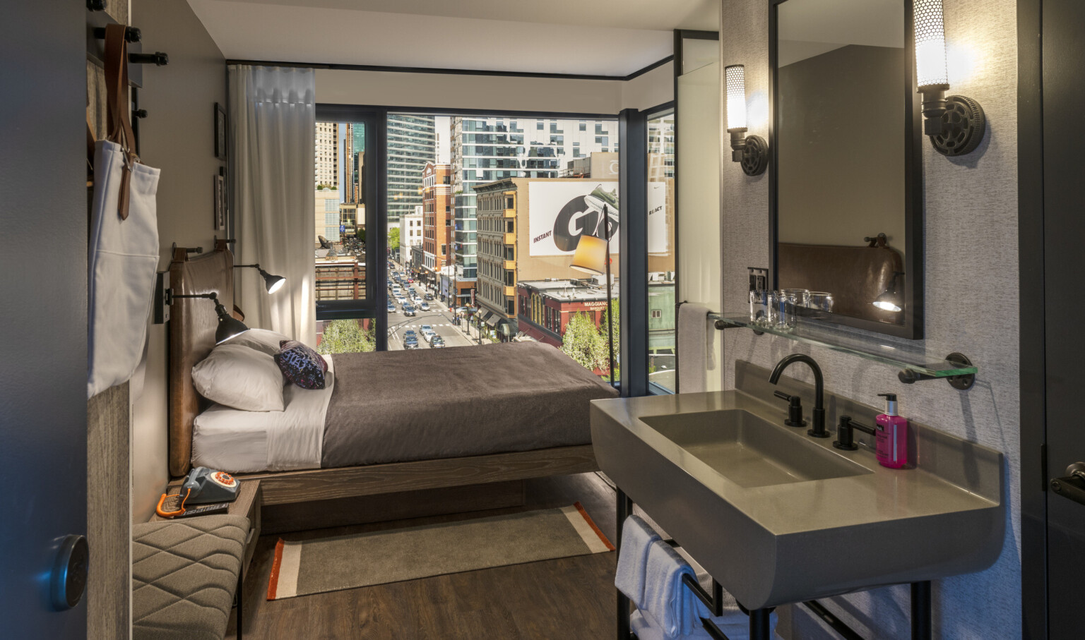 View of guest room from grey sink, looking towards bed and large corner window. White room with grey around sink and mirror