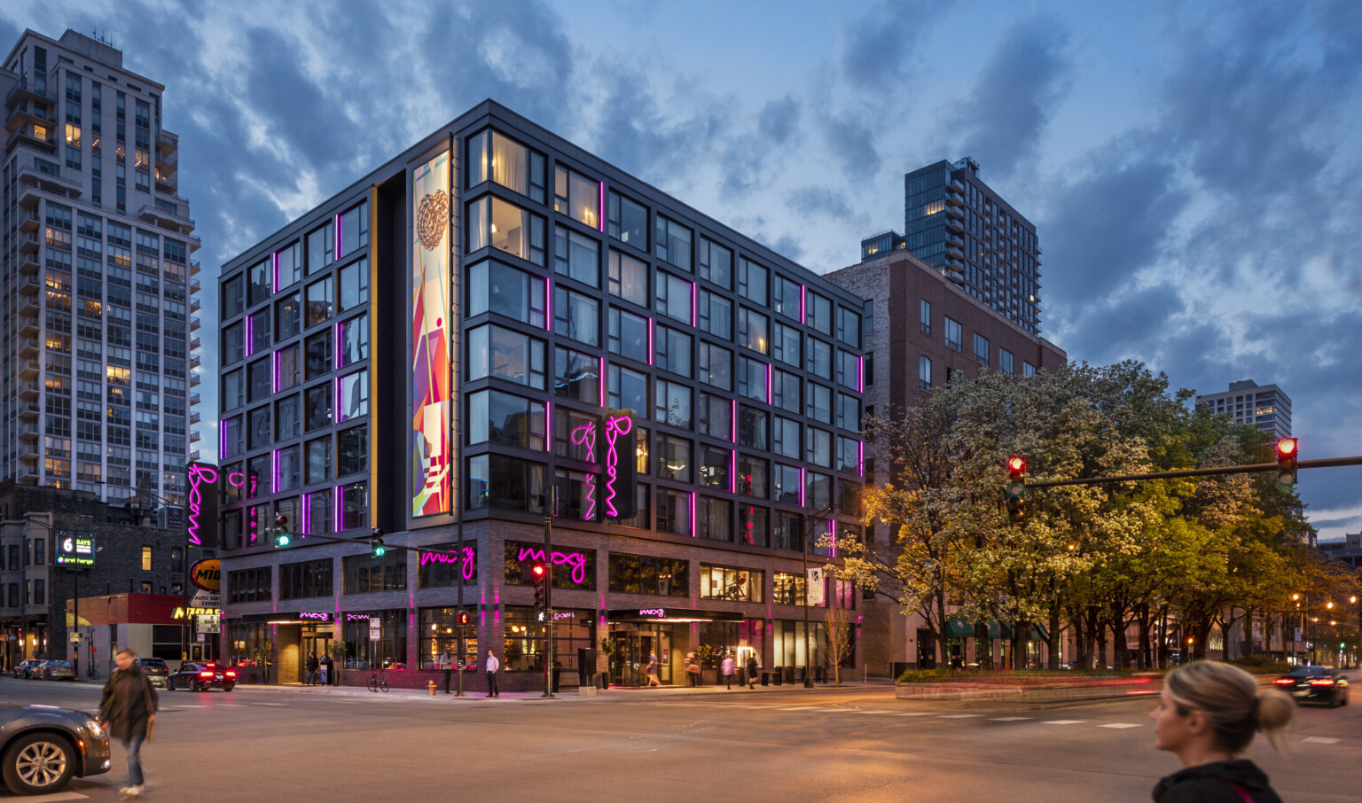 Corner view of the Moxy hotel with illuminated mural and pink lights at various windows. 2 brick stories, exposed steel above