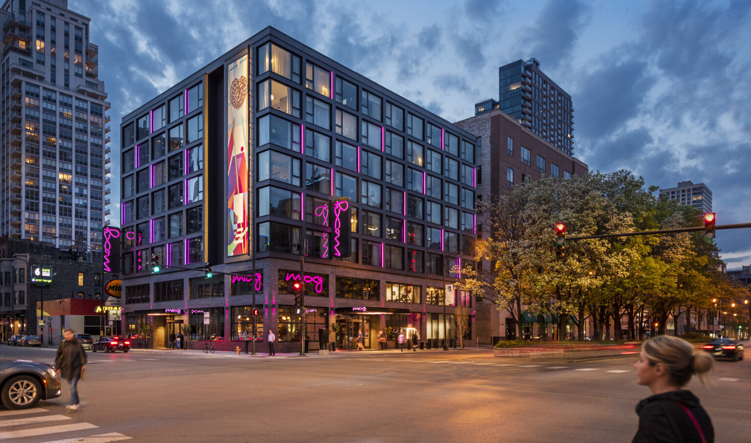 Corner view of the Moxy hotel with illuminated mural and pink lights at various windows. 2 brick stories, exposed steel above