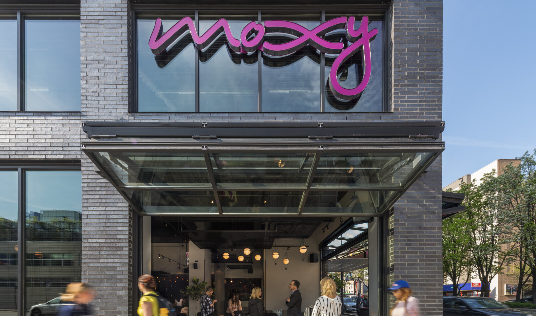1st floor corner of building with window below pink Moxy sign opened up and out above sidewalk looking into dining area