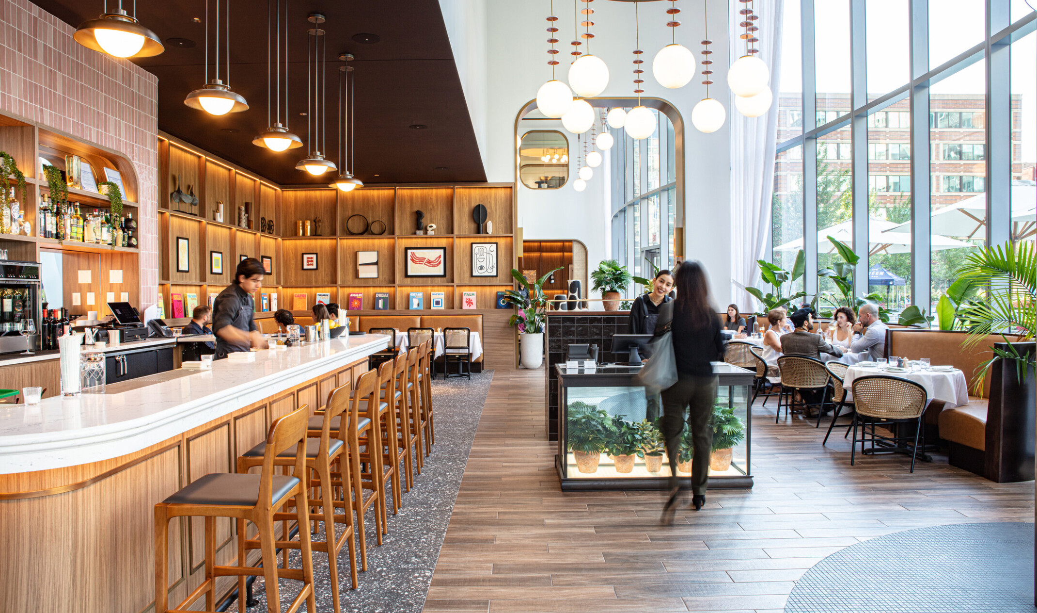 Modern coffee shop with long counter lined with bar height stools, wooden shelves, and bench seating lining floor to ceiling windows
