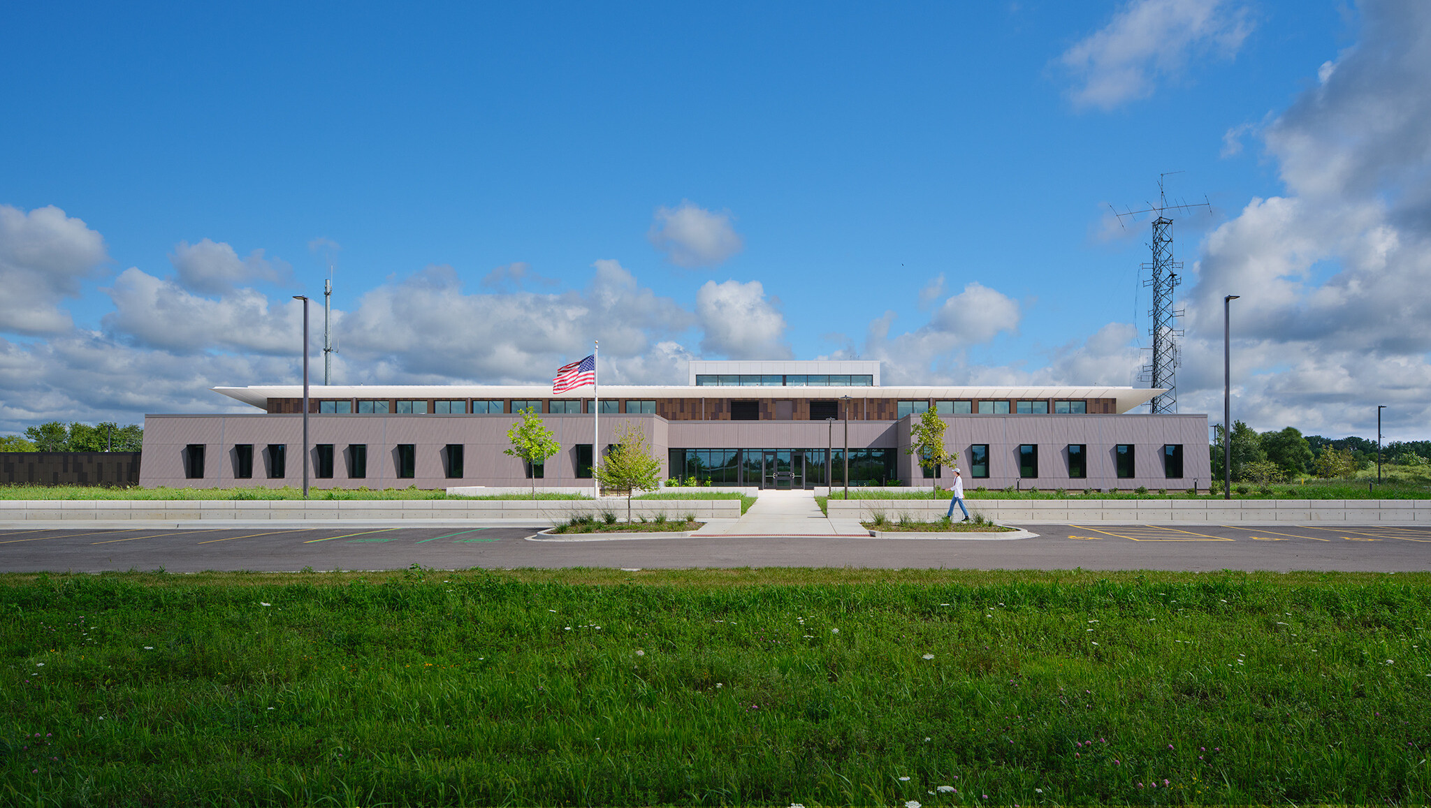 Rendering of a one-story brick building with white butterfly wing roof and tall communications antenna on a grassy site with wide sidewalks around the perimeter and a main drive that is protected by bollards at the entrance.