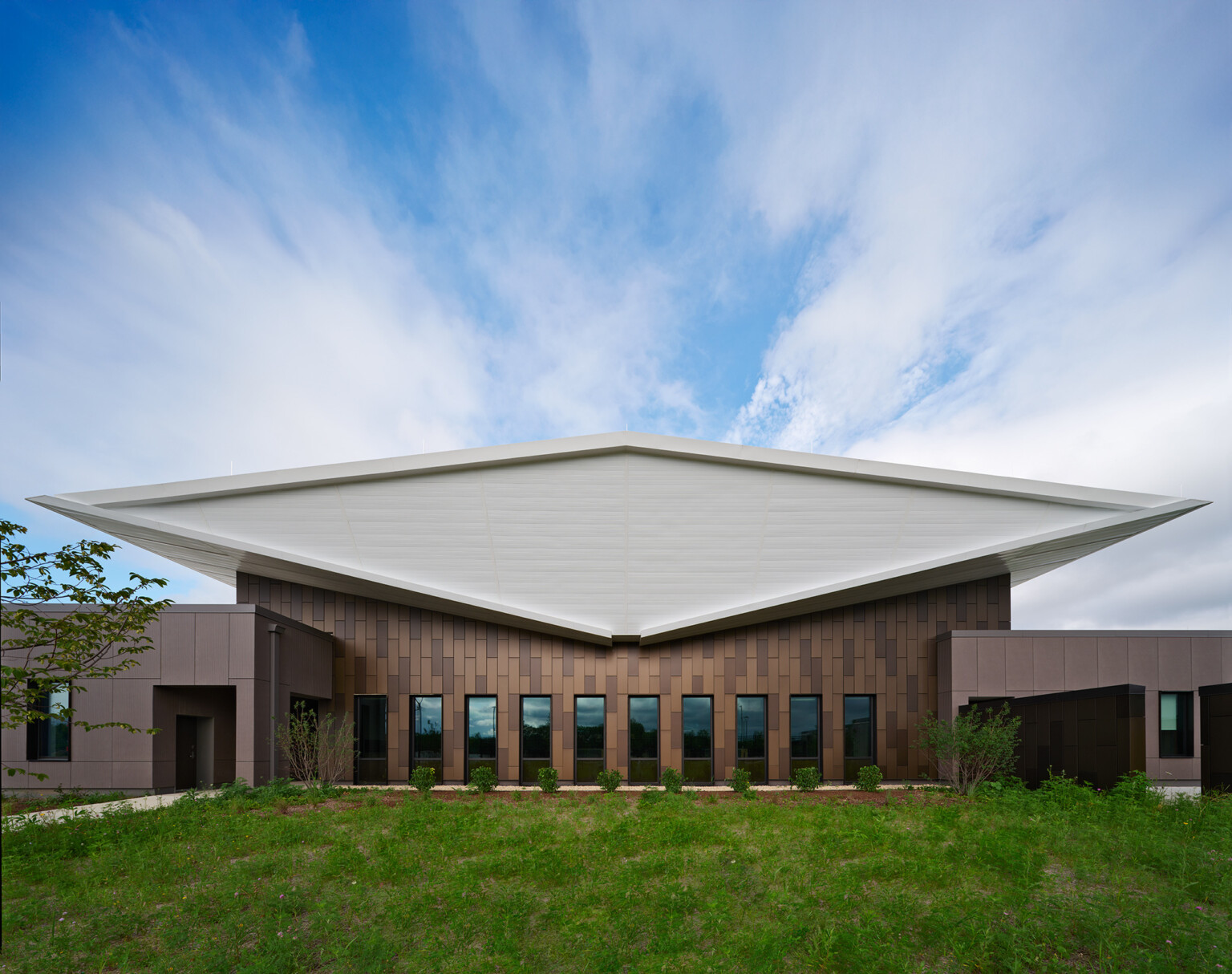 Exterior of lake county regional operations and communications facility; Head-on view of the building’s exterior end shows the white double-butterfly roof as a diamond-shaped volume that floats above the dark brown textured building exterior that is pieced by narrow vertical windows.