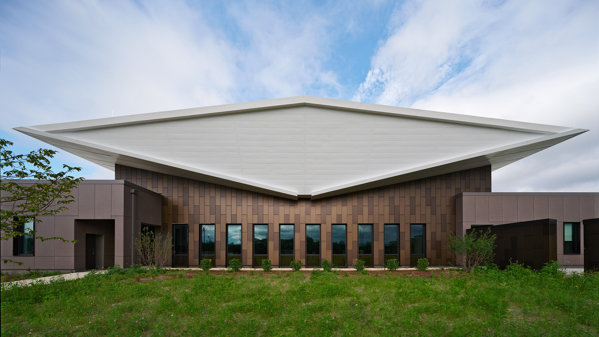 Exterior of lake county regional operations and communications facility; Head-on view of the building’s exterior end shows the white double-butterfly roof as a diamond-shaped volume that floats above the dark brown textured building exterior that is pieced by narrow vertical windows.