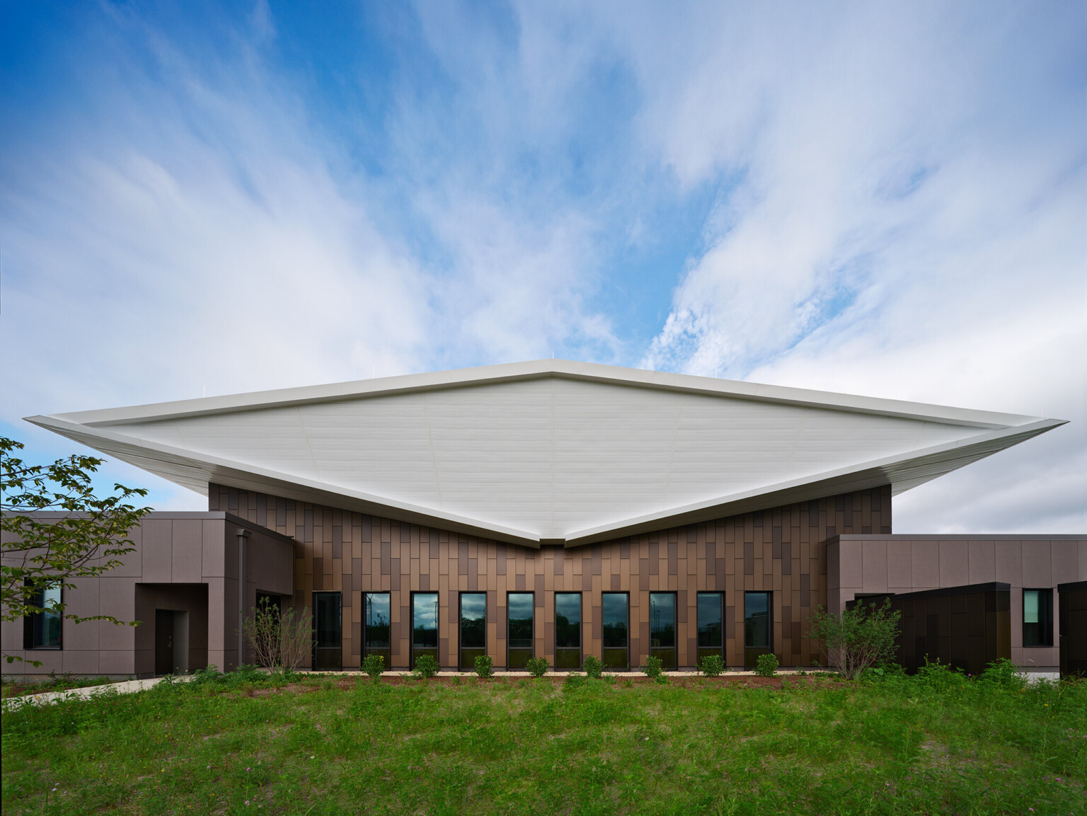 Exterior of lake county regional operations and communications facility; Head-on view of the building’s exterior end shows the white double-butterfly roof as a diamond-shaped volume that floats above the dark brown textured building exterior that is pieced by narrow vertical windows.