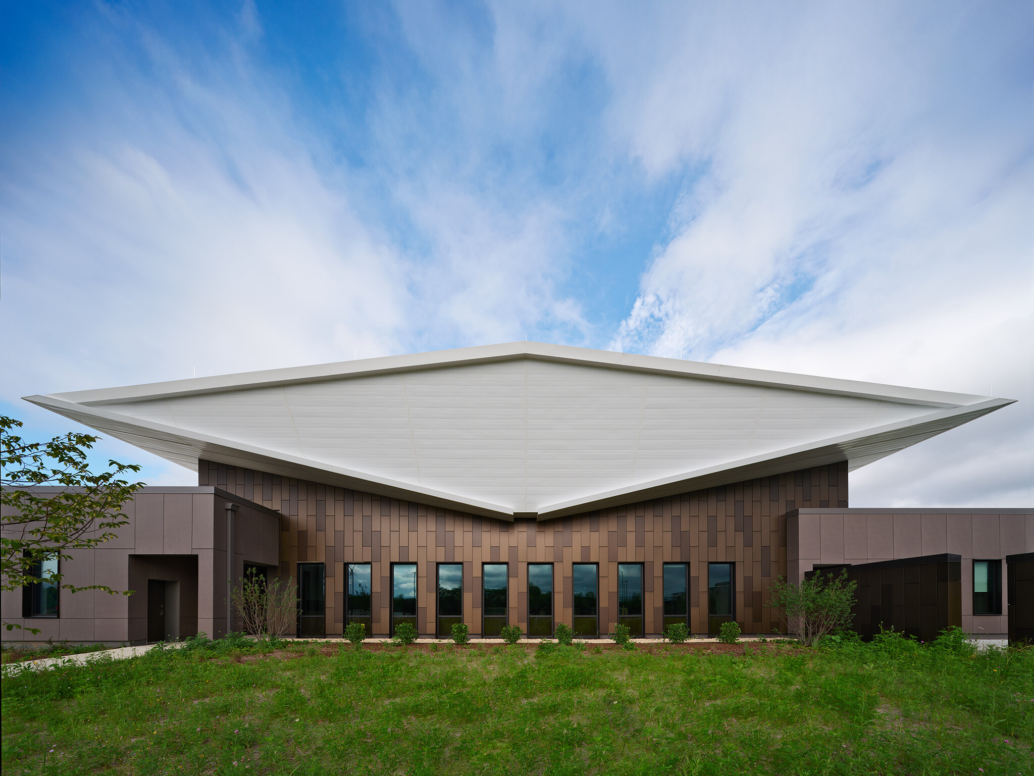 Exterior of lake county regional operations and communications facility; Head-on view of the building’s exterior end shows the white double-butterfly roof as a diamond-shaped volume that floats above the dark brown textured building exterior that is pieced by narrow vertical windows.