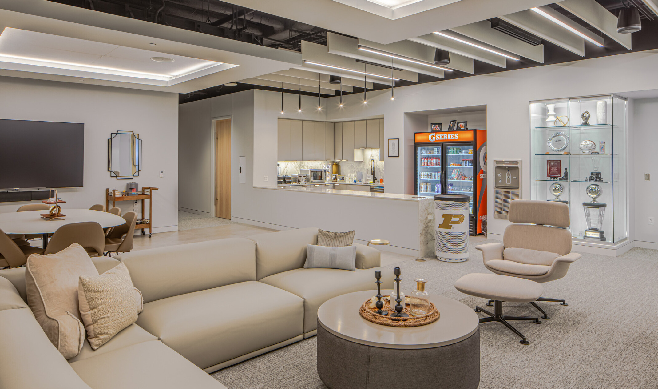 Large lounge with display case, tan L shaped couch, seating area in front of a TV, and kitchen area in the back