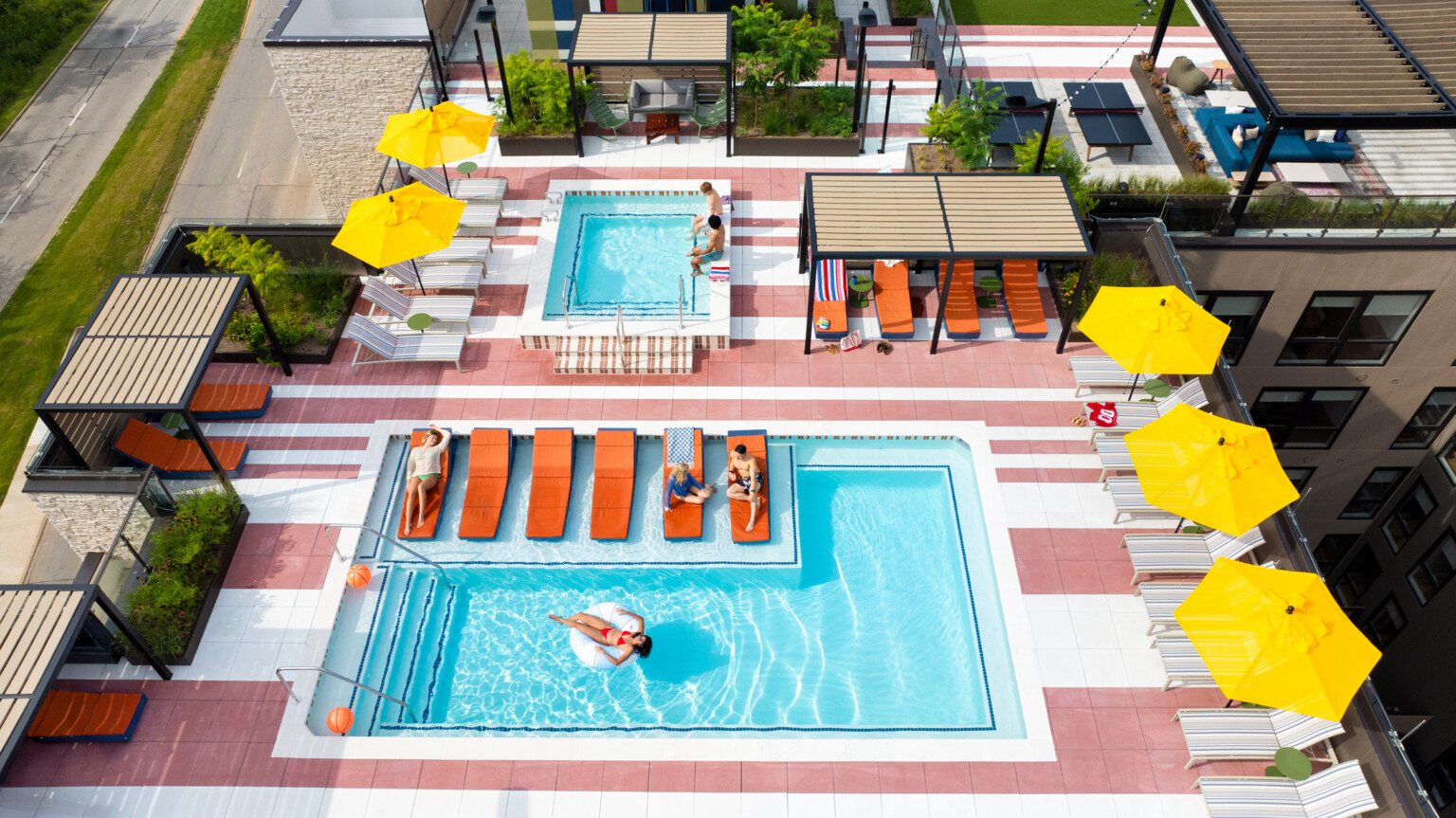 Hub Bloomington off campus student housing; Aerial view of resort-style rooftop pool with cabanas, umbrellas, and students relaxing and swimming.