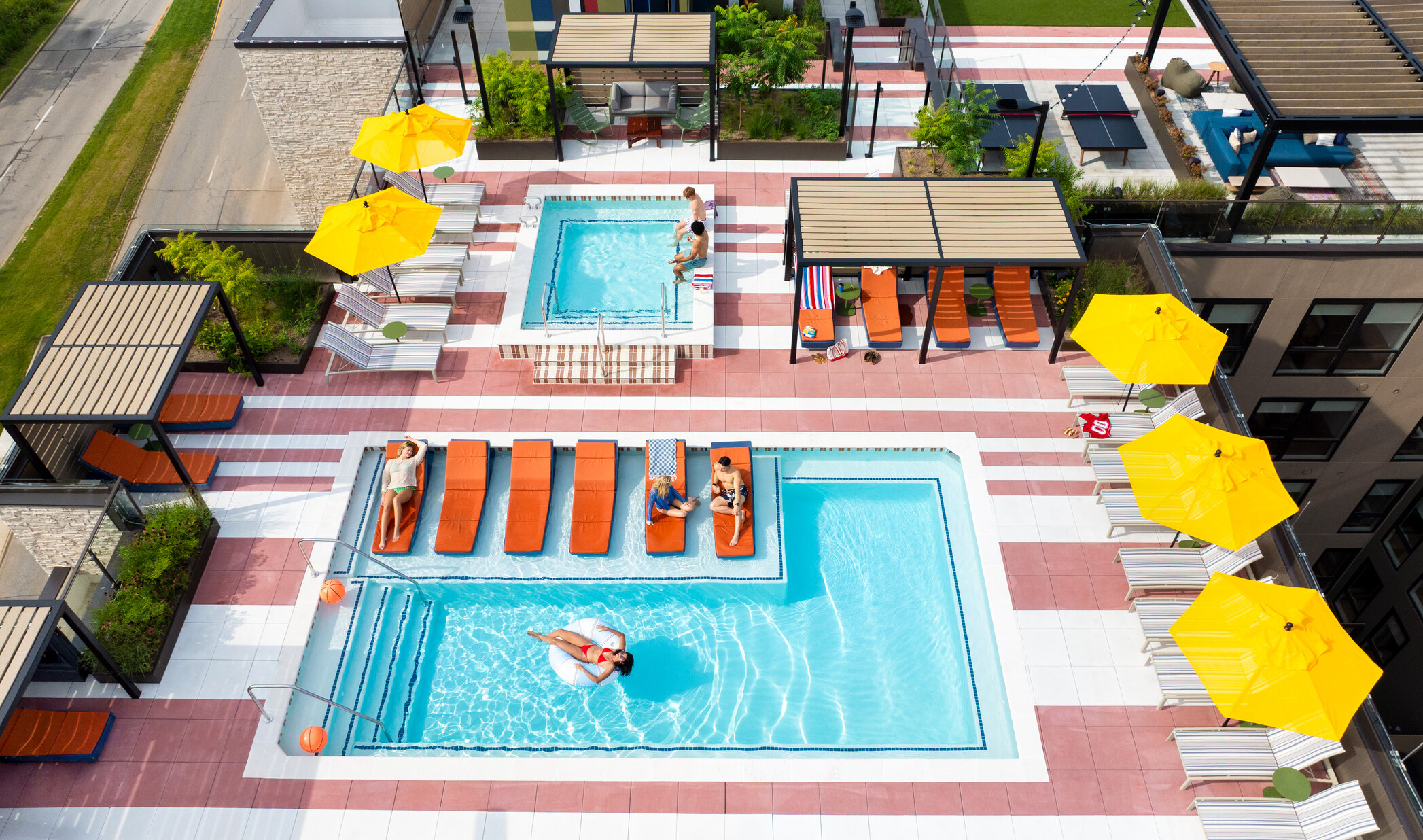 Hub Bloomington off campus student housing; Aerial view of resort-style rooftop pool with cabanas, umbrellas, and students relaxing and swimming.