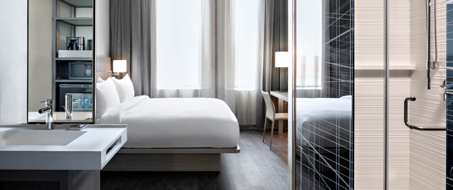 Guest room with white bed partially separated from kitchenette by translucent and metal wall right. Sink with mirror left following Marriott brand architecture