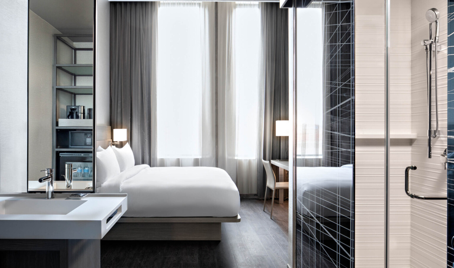 Guest room with white bed partially separated from kitchenette by translucent and metal wall right. Sink with mirror left