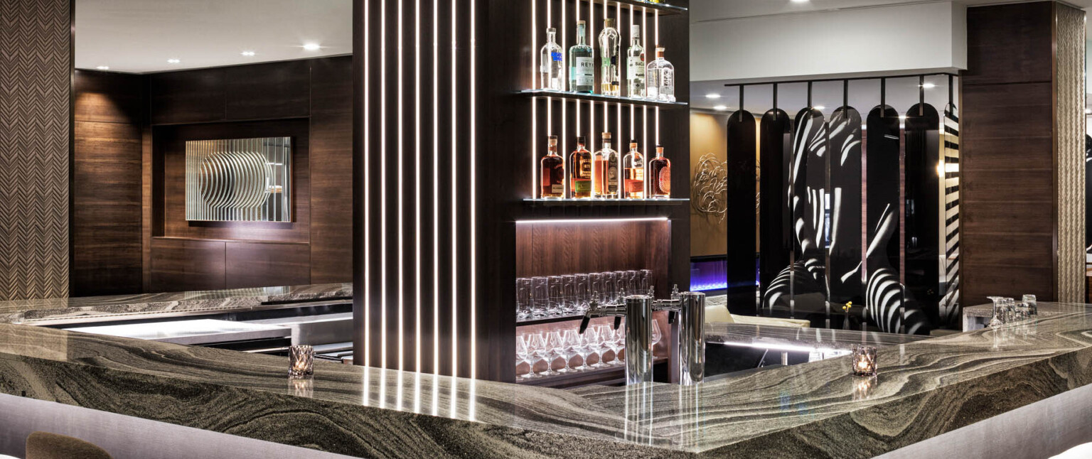 Rounded bar with dark marble counter and central wood pillar with shelving and illuminated recessed strips in wood panel room following Marriott brand architecture