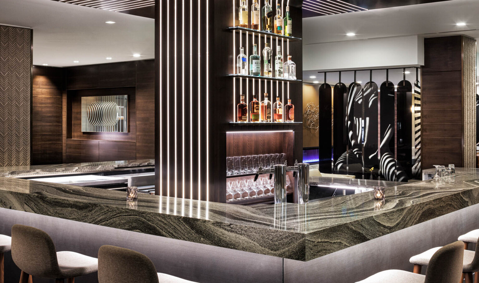 Rounded bar with dark marble counter and central wood pillar with shelving and illuminated recessed strips in wood panel room