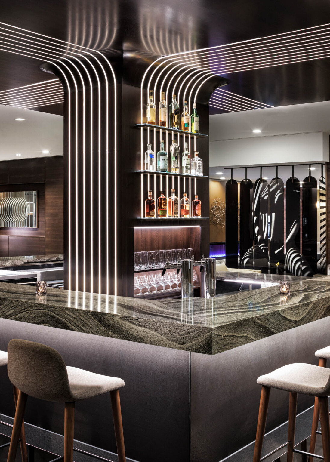 Wider view of bar, strips of light curve up the wall onto ceiling detail above. Grey cushioned stools line black base of bar